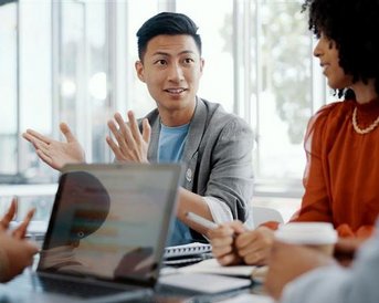 Ein Mann spricht lebhaft in einem Besprechungsraum, während andere Teilnehmer aufmerksam zuhören. Laptops und Notizen sind auf dem Tisch verteilt.