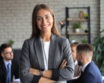 Eine lächelnde Frau in einem grauen Blazer steht im Vordergrund, während im Hintergrund mehrere Personen an einem Tisch diskutieren.