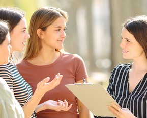 Four women are standing outside and discussing. One woman is holding a clipboard and speaking, while the others listen attentively.