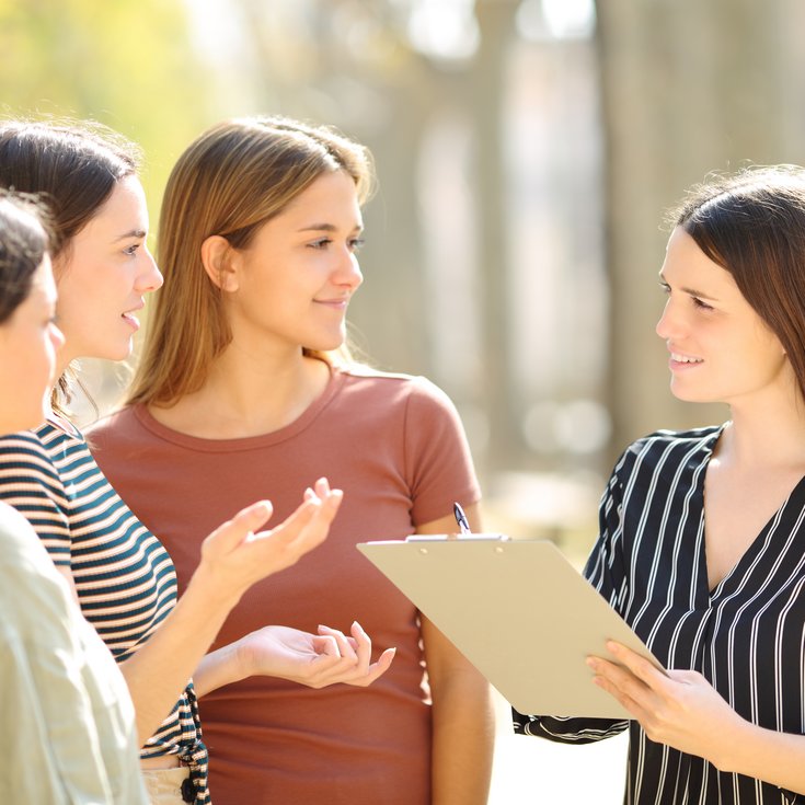 Four women are standing outside and discussing. One woman is holding a clipboard and speaking, while the others listen attentively.