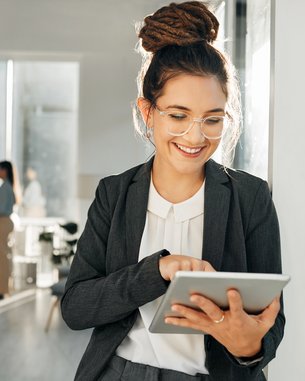 Eine lächelnde Frau in einem Anzug nutzt ein Tablet, während sie in einem modernen Büro steht. Im Hintergrund sind mehrere Personen zu sehen.