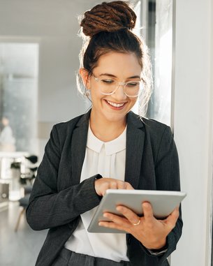 Eine lächelnde Frau in einem Anzug nutzt ein Tablet, während sie in einem modernen Büro steht. Im Hintergrund sind mehrere Personen zu sehen.