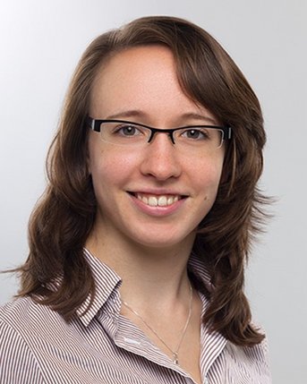 Eine junge Frau mit schulterlangen, lockigen Haaren und Brille lächelt freundlich. Sie trägt ein gestreiftes Hemd und hat einen neutralen Hintergrund.