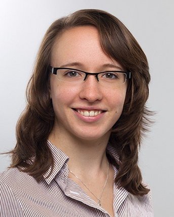 Eine junge Frau mit schulterlangen, lockigen Haaren und Brille lächelt freundlich. Sie trägt ein gestreiftes Hemd und hat einen neutralen Hintergrund.