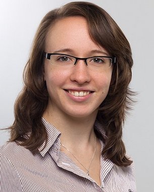 Eine junge Frau mit schulterlangen, lockigen Haaren und Brille lächelt freundlich. Sie trägt ein gestreiftes Hemd und hat einen neutralen Hintergrund.