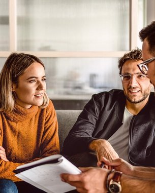 Zwei Personen sitzen auf einem Sofa und unterhalten sich, während eine dritte Person Notizen macht. Die Frau trägt einen orangefarbenen Pullover, der Mann neben ihr hat eine schwarze Jacke an. Der dritte Mann trägt eine Brille.