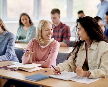Three young women are sitting at a table in a classroom, chatting and taking notes.