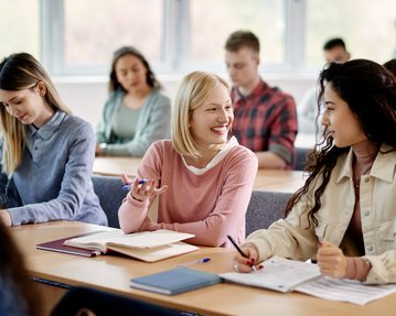 Three young women are sitting at a table in a classroom, chatting and taking notes.