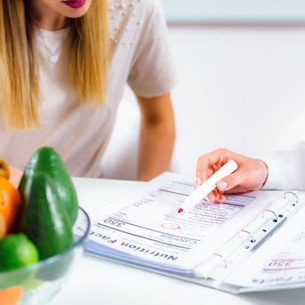 Eine Person zeigt auf ein Dokument in einem Ordner, während eine andere Person mit langen Haaren und einem weißen Oberteil zuschaut. Eine Schüssel mit Obst steht auf dem Tisch.