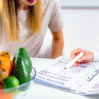 Eine Person zeigt auf ein Dokument in einem Ordner, während eine andere Person mit langen Haaren und einem weißen Oberteil zuschaut. Eine Schüssel mit Obst steht auf dem Tisch.