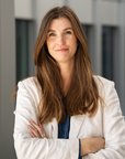 Eine Frau mit langen, braunen Haaren und einem weißen Blazer steht mit verschränkten Armen vor einem modernen Gebäude.