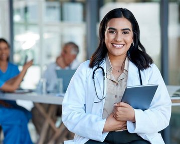 Eine lächelnde Ärztin in einem weißen Kittel hält ein Tablet in der Hand, während im Hintergrund Kollegen in medizinischer Kleidung diskutieren.