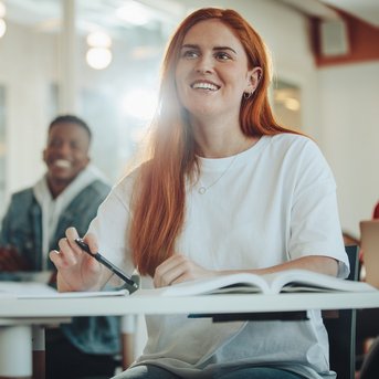 Eine Gruppe von Studierenden sitzt in einem Klassenzimmer. Eine Frau mit langen roten Haaren lächelt, während sie Notizen macht. Im Hintergrund lachen andere Studierende.