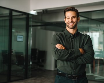 Ein junger Mann mit einem breiten Lächeln steht in einem modernen Büro, die Arme vor der Brust verschränkt, in einem dunkelgrünen Hemd.