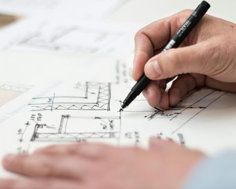 Eine Hand zeichnet technische Pläne mit einem schwarzen Stift auf Papier. Die Skizzen zeigen verschiedene Bau- und Konstruktionsdetails.
