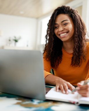 Eine Frau mit lockigem Haar sitzt an einem Schreibtisch und lächelt, während sie an einem Laptop arbeitet. Sie hält einen Stift in der Hand und notiert etwas in einem Notizbuch. Pflanzen und Schreibmaterial sind sichtbar.