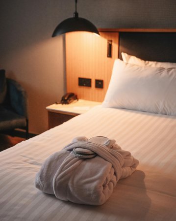 A folded bathrobe lies on a bed with striped bedding, next to it a lamp and a dresser with power outlets.