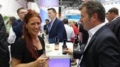 Eine Frau mit rotem Haar hält ein Glas, während sie mit einem Mann in Anzug spricht. Im Hintergrund sind weitere Personen und Stände zu sehen.