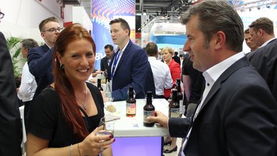 Eine Frau mit rotem Haar hält ein Glas, während sie mit einem Mann in Anzug spricht. Im Hintergrund sind weitere Personen und Stände zu sehen.