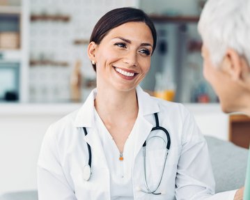 Eine Ärztin in weißem Kittel lächelt und spricht mit einer älteren Patientin in einem hellen, modernen Raum.