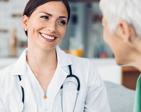 Eine Ärztin in weißem Kittel lächelt und spricht mit einer älteren Patientin in einem hellen, modernen Raum.