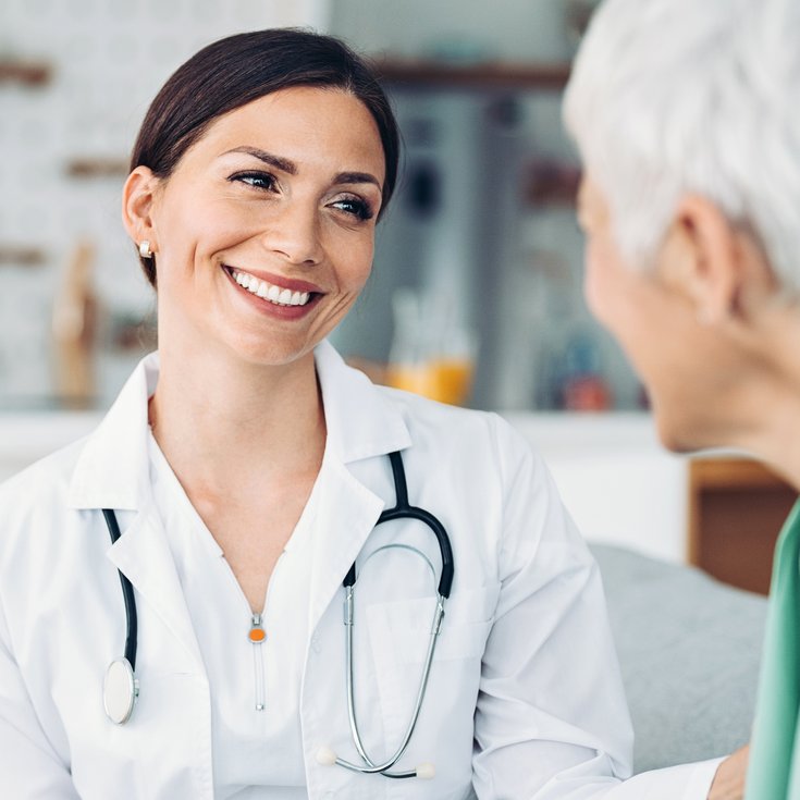 Eine Ärztin in weißem Kittel lächelt und spricht mit einer älteren Patientin in einem hellen, modernen Raum.