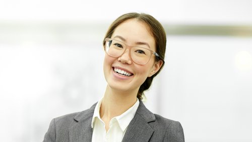 Eine lächelnde Frau mit Brille trägt einen grauen Anzug und ein weißes Hemd. Sie steht in einem modernen Büro.