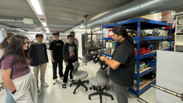 A group of five people is watching a man demonstrate a device in a lab or workshop. In the background, there are shelves filled with tools.
