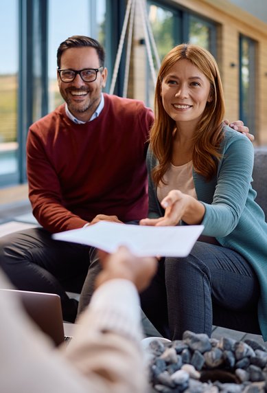 Ein Paar sitzt auf einer Couch und lächelt, während sie ein Dokument von einer Person entgegennehmen, die ihnen gegenüber sitzt.