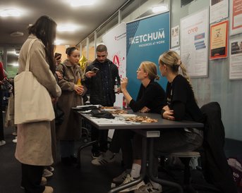 A group of people is standing around a table while two women are offering information and materials. In the background, posters and banners are visible.