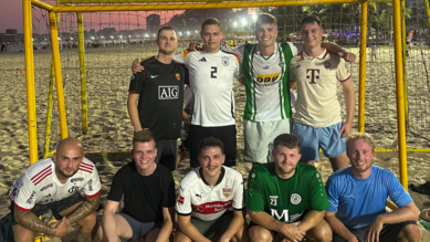 Neun Männer posieren in Sportkleidung vor einem Fußballtor am Strand, lächelnd und in zwei Reihen angeordnet.