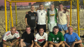 Neun Männer posieren in Sportkleidung vor einem Fußballtor am Strand, lächelnd und in zwei Reihen angeordnet.