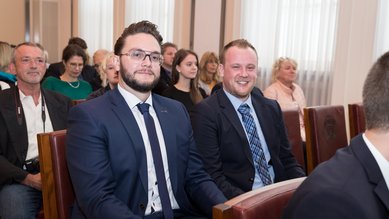 Zwei Männer in Anzügen sitzen in einer Reihe von Stühlen, umgeben von einer Gruppe von Menschen in einem formellen Veranstaltungsraum.