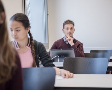 A young man sits in a classroom, thoughtfully resting his chin on his hand. In the foreground, a female student is looking at a laptop. Several empty desks are visible in the background.
