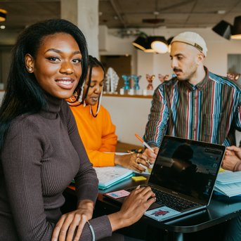 Eine Frau mit langen, schwarzen Haaren sitzt an einem Tisch und lächelt, während sie auf einen Laptop schaut. Um sie herum arbeiten drei weitere Personen an Notizen und Projekten. Der Raum ist modern und hell.