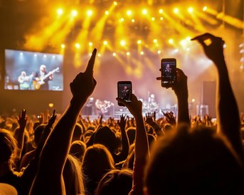 A crowd at a concert filming the performance with their phones. Bright lights and a band on stage are visible.