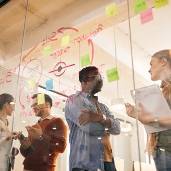 Vier Personen diskutieren vor einer Glaswand mit bunten Haftnotizen und Diagrammen, während sie Ideen austauschen und Notizen machen.