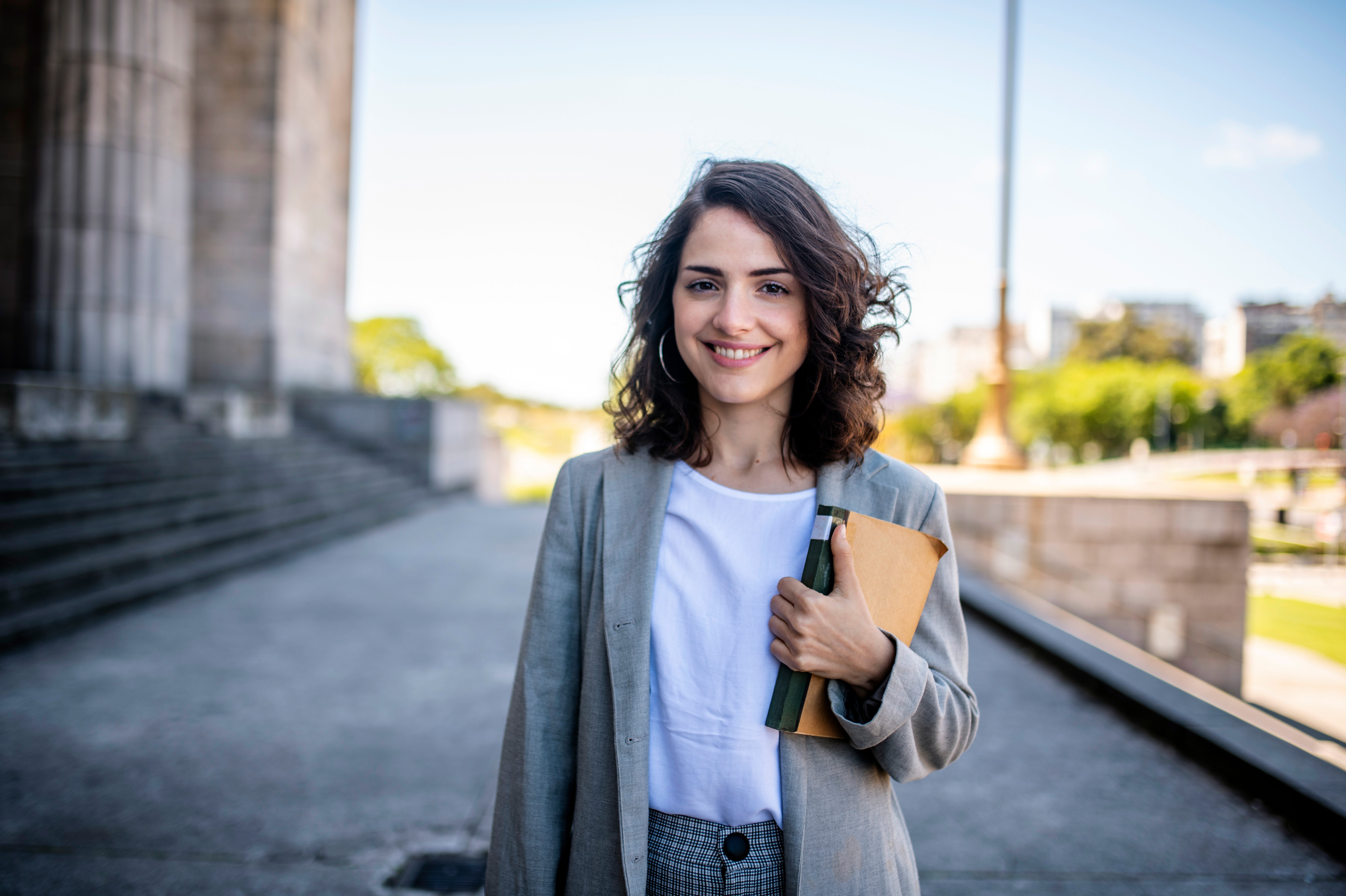 Junge Frau mit lockigem Haar trägt einen grauen Blazer und hält ein Notizbuch sowie einen Stift. Sie steht im Freien vor einer monumentalen Struktur und lächelt freundlich in die Kamera.