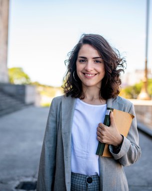 Junge Frau mit lockigem Haar trägt einen grauen Blazer und hält ein Notizbuch sowie einen Stift. Sie steht im Freien vor einer monumentalen Struktur und lächelt freundlich in die Kamera.