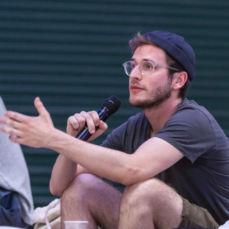Ein junger Mann mit Brille und Mütze spricht leidenschaftlich in ein Mikrofon, während er auf einem Stuhl sitzt und gestikuliert.