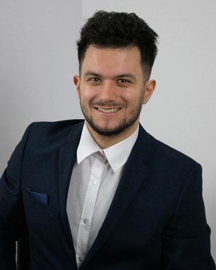 A young man in a suit smiles at the camera, wearing a white shirt and a dark blazer.