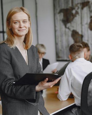 Eine Frau in einem Anzug hält ein Notizbuch und lächelt, während sie in einem modernen Büroraum steht. Im Hintergrund sind mehrere Personen in einem Meeting zu sehen.