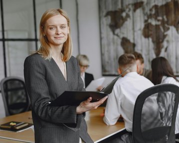Eine Frau in einem Anzug hält ein Notizbuch und lächelt, während sie in einem modernen Büroraum steht. Im Hintergrund sind mehrere Personen in einem Meeting zu sehen.