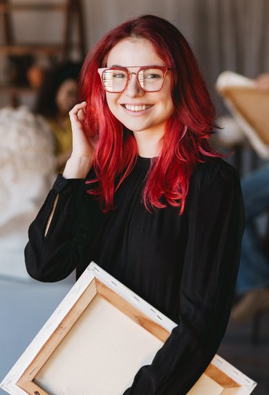 Eine lächelnde Frau mit roten Haaren und Brille hält eine Leinwand in einem Kunststudio, während im Hintergrund andere Personen zeichnen.