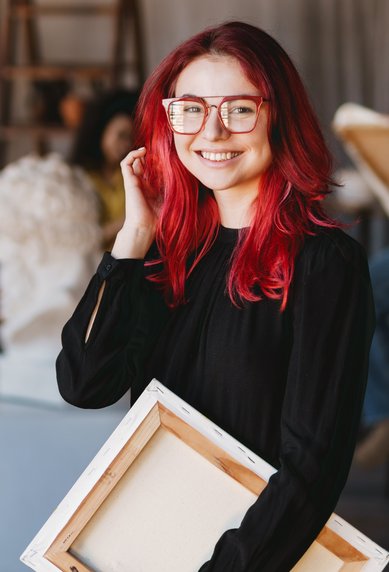 A smiling woman with red hair and glasses holds a canvas in an art studio, while others are drawing in the background.