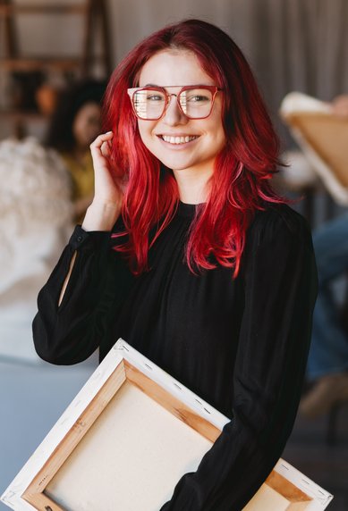 Eine lächelnde Frau mit roten Haaren und Brille hält eine Leinwand in einem Kunststudio, während im Hintergrund andere Personen zeichnen.