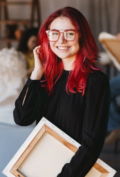 A smiling woman with red hair and glasses holds a canvas in an art studio, while others are drawing in the background.
