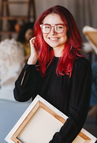 Eine lächelnde Frau mit roten Haaren und Brille hält eine Leinwand in einem Kunststudio, während im Hintergrund andere Personen zeichnen.