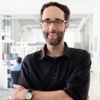 Ein Mann mit Brille und Bart steht mit verschränkten Armen in einem modernen Büro. Er trägt ein schwarzes Hemd und eine Armbanduhr.