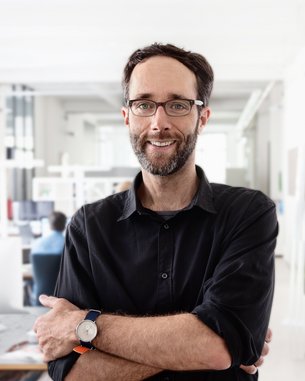 A man with glasses and a beard stands with his arms crossed in a modern office. He is wearing a black shirt and a wristwatch.